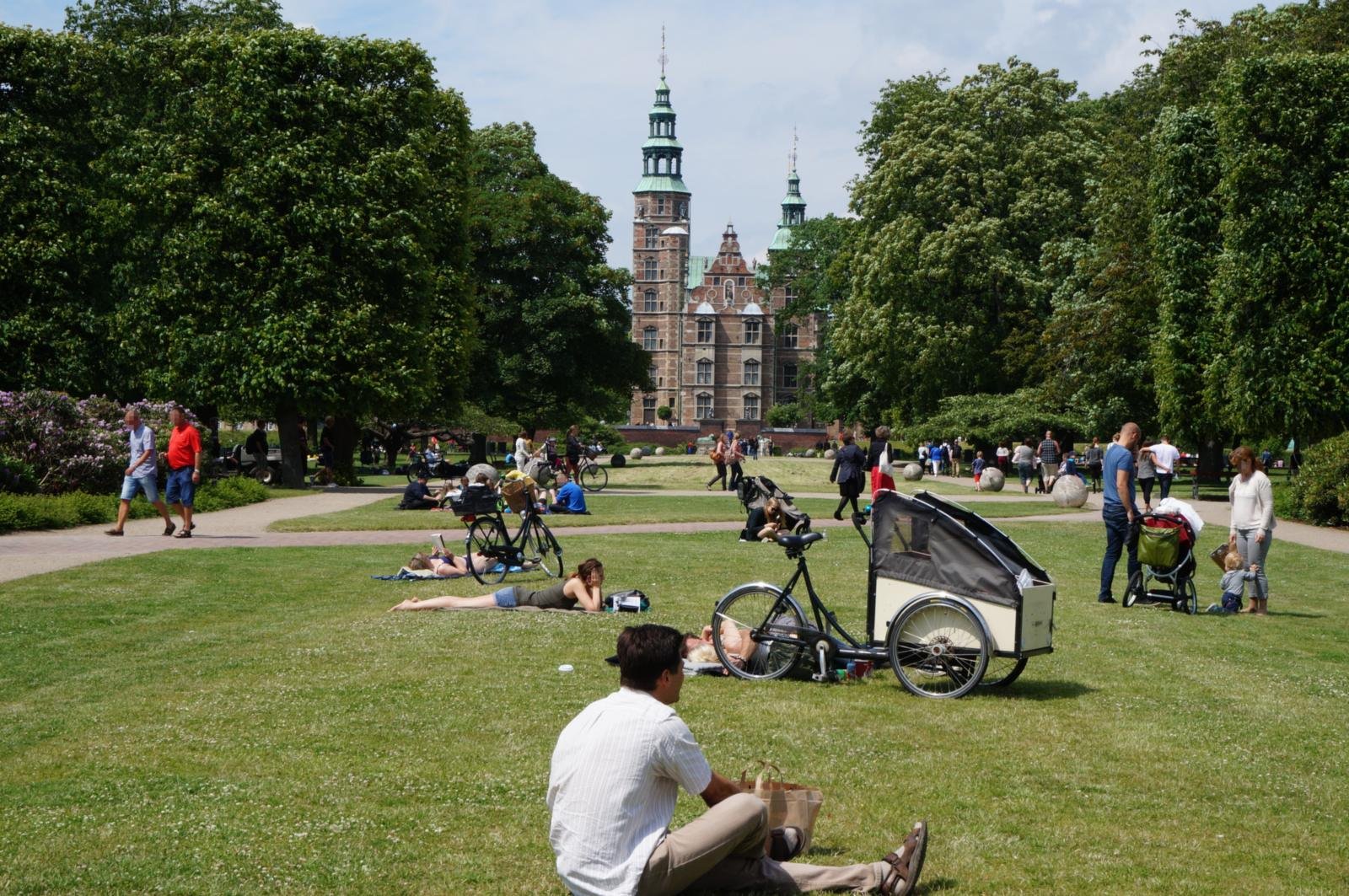 Kongens Have on a summer day: people on the lawn and a cargo bike in front of the green copper towers of Rosenborg Castle.